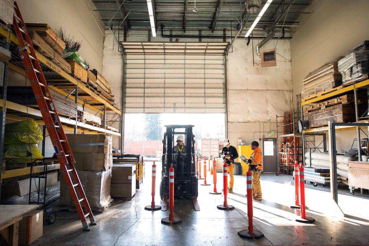 1456969202 A multiracial woman worker operates a forklift, her coworkers reviewing paperwork for the day in a warehouse setting, wearing safety gear.   Confident safe workers in their work place.
